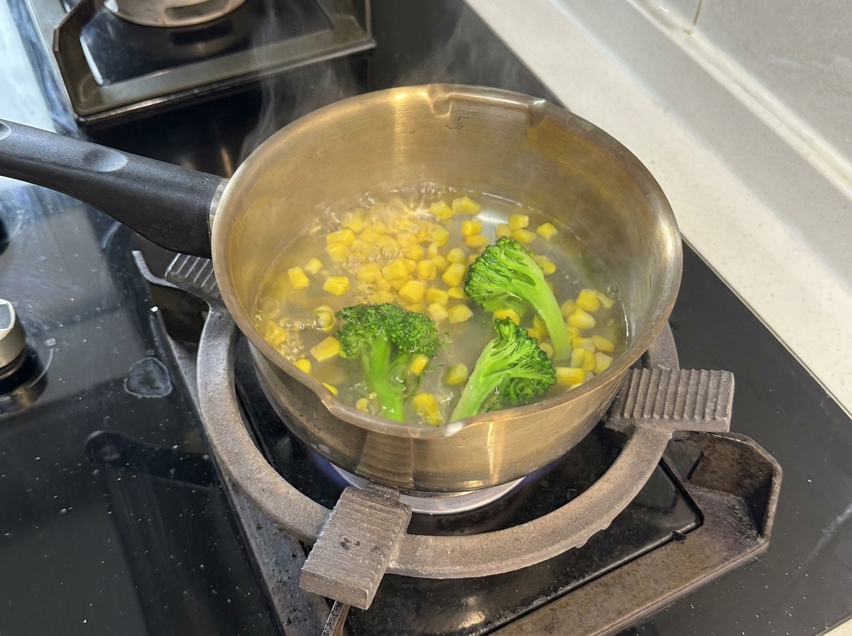 A stainless steel pot on a stovetop filled with boiling water, broccoli florets, and yellow corn kernels.