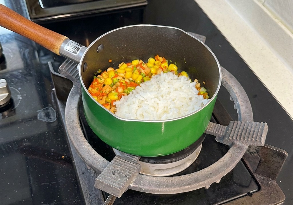 A scoop of white steamed rice sits atop a colorful mixture of stir-fried salmon cubes, corn, carrots, and broccoli in a green saucepan.