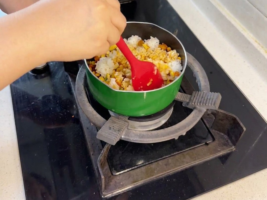 A person using a red spatula to mix white steamed rice with stir-fried salmon and vegetables in a green pan on a stovetop.
