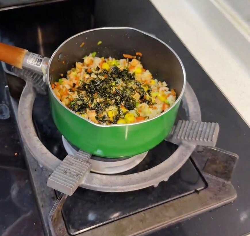 Dark green seaweed flakes being sprinkled from a small glass jar onto a pan of freshly mixed salmon and vegetable rice.
