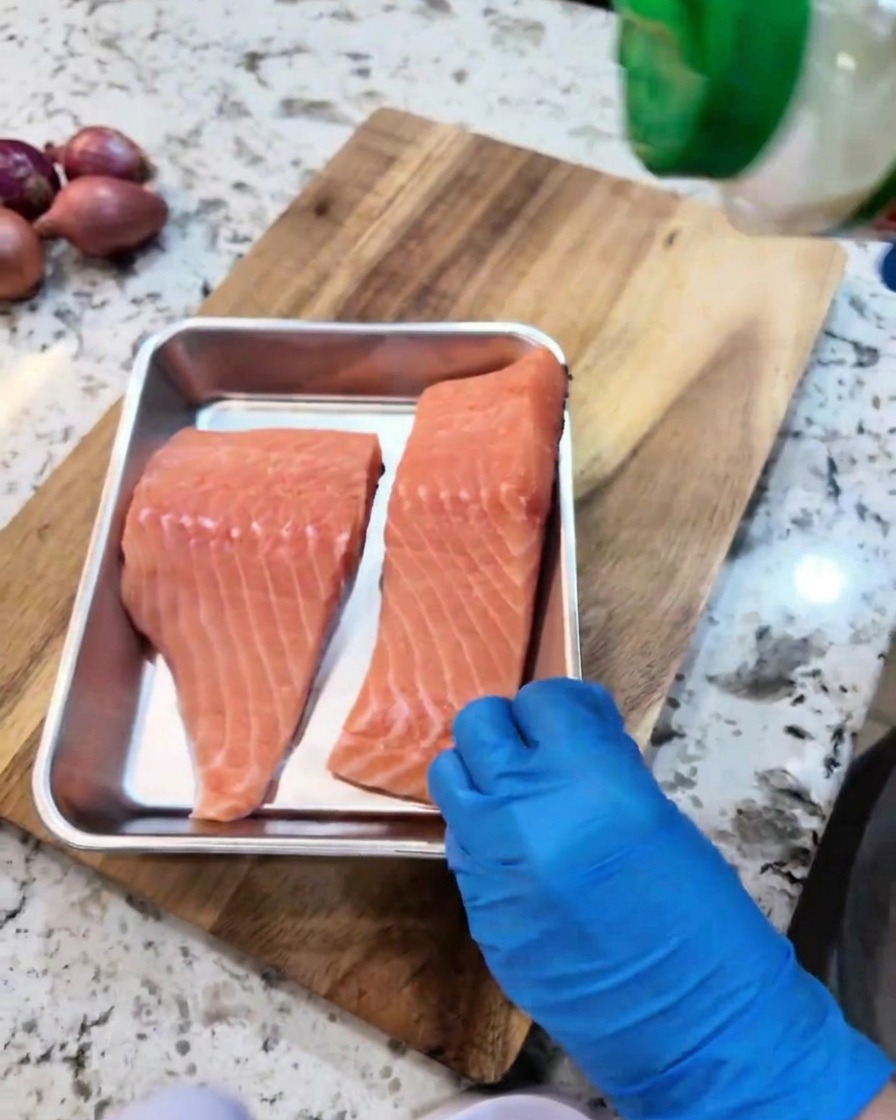 Raw salmon fillets being seasoned with garlic powder and black pepper in a metal tray.
