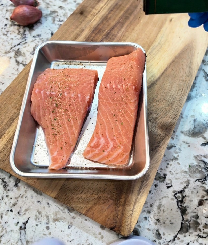 A hand seasoning raw salmon fillets in a metal tray with a sprinkle of black pepper and garlic powder.