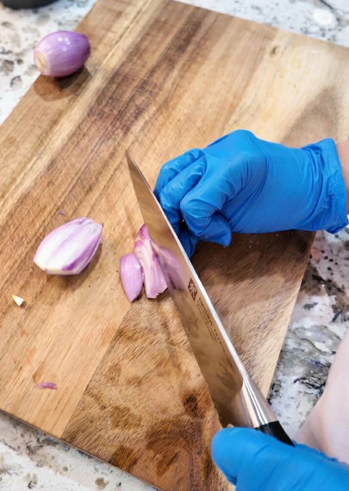 A kitchen knife slicing purple shallots into thin rounds on a wooden board.