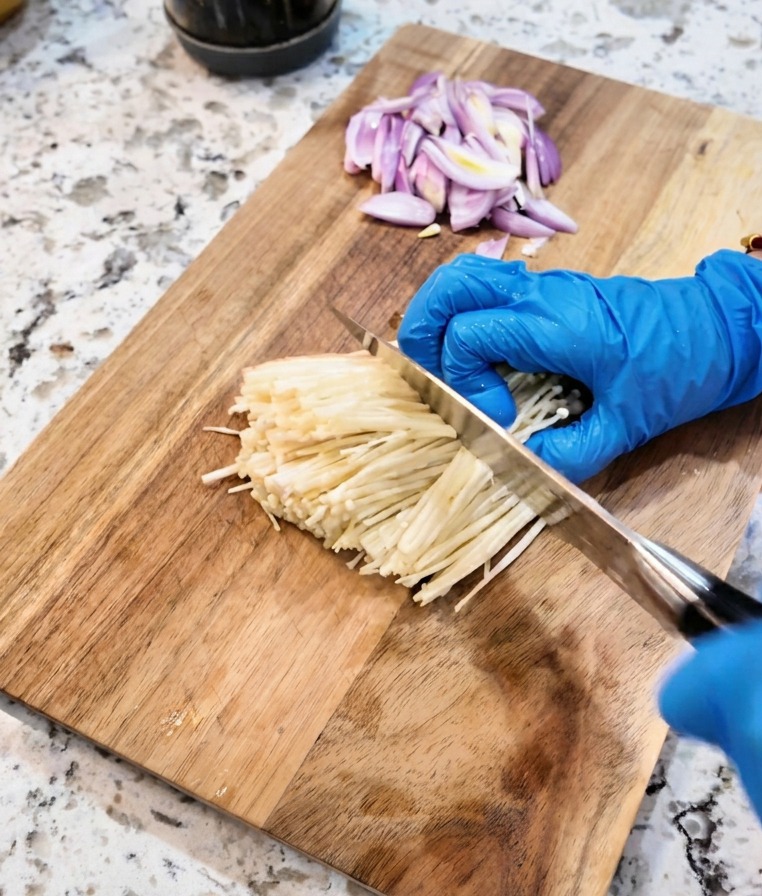A knife cutting off the base of a bunch of white enoki mushrooms on a cutting board.