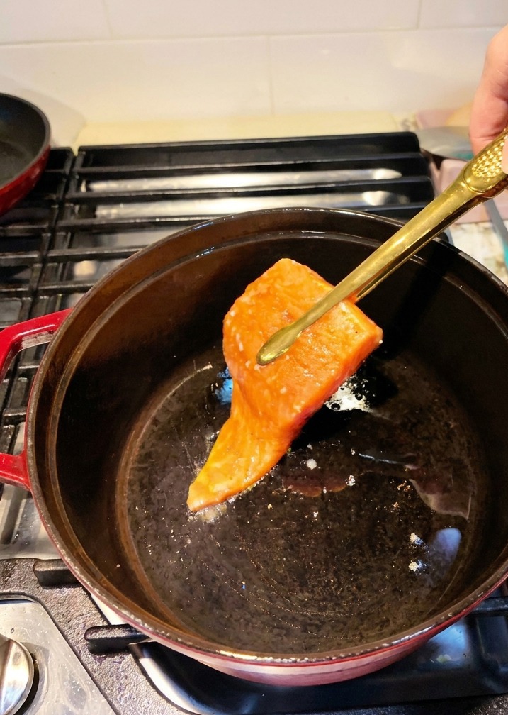 Marinated salmon fillets being placed into a preheated black cast iron pot using kitchen tongs.
