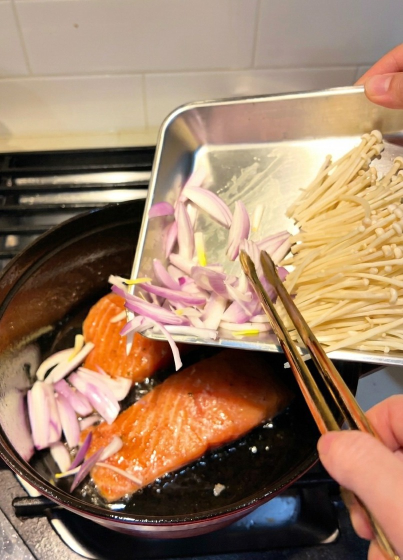 Sliced shallots being added to a pot where salmon fillets are currently searing in oil.