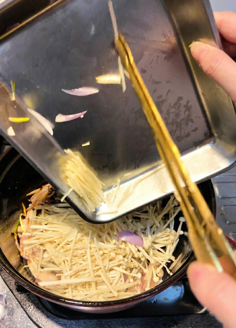 Enoki mushrooms being added to a pot to be stir-fried with sautéed shallots.