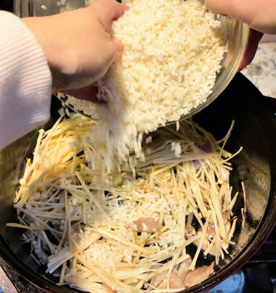 Two cups of white rice being poured from a bowl into a pot containing mushrooms.