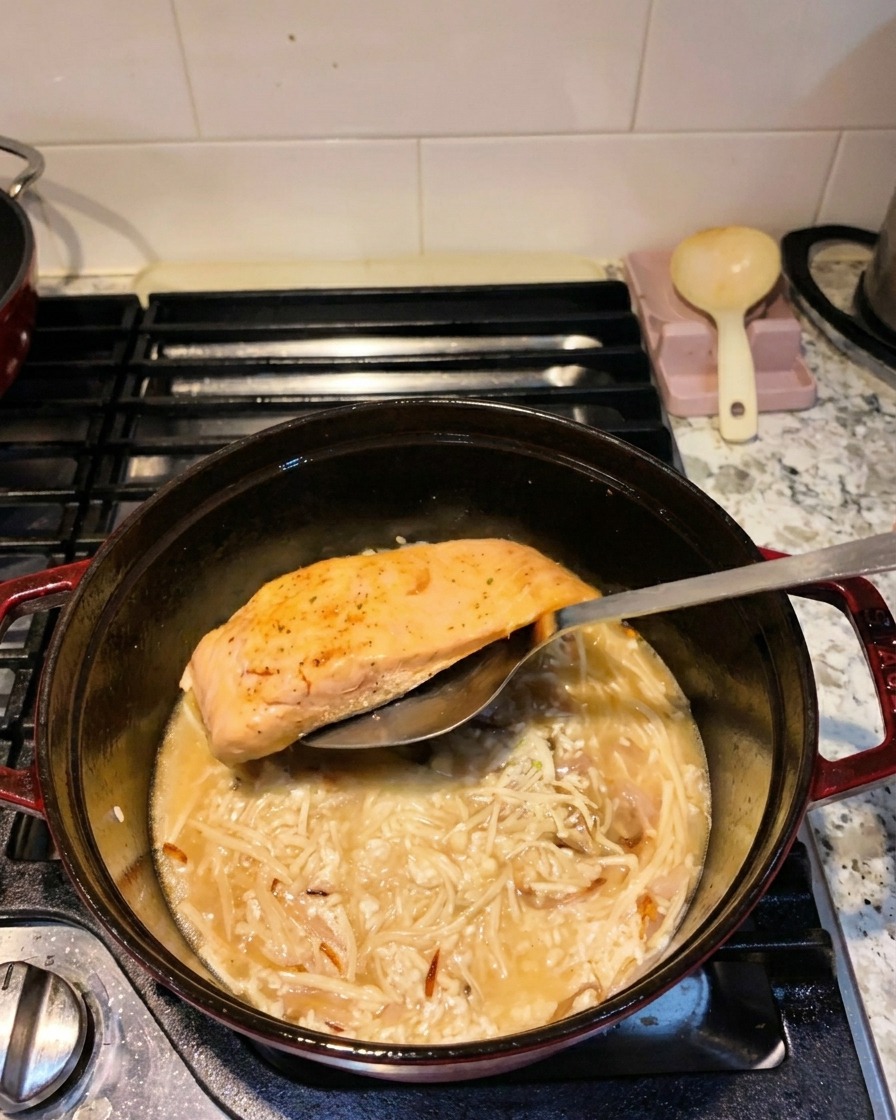 A metal spatula lowering a seared salmon fillet into a pot containing rice, enoki mushrooms, and cooking liquid.
