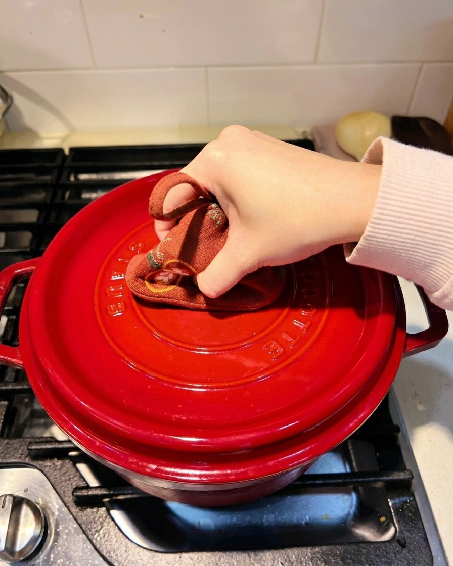 A hand using a patterned pot holder to press down the lid of a red cast-iron pot on a stovetop.