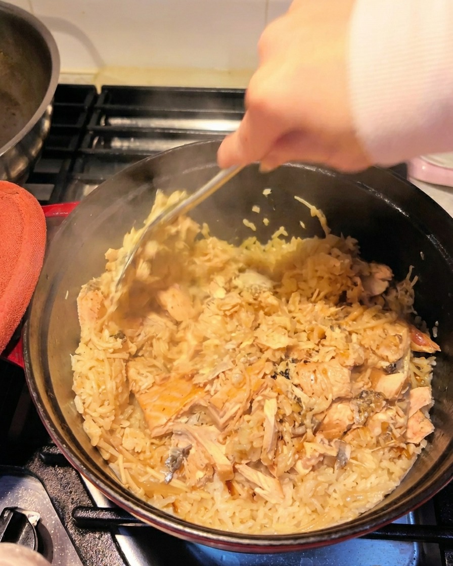 A hand using a metal spatula to break up cooked salmon fillets and mix them into the seasoned rice inside the pot.