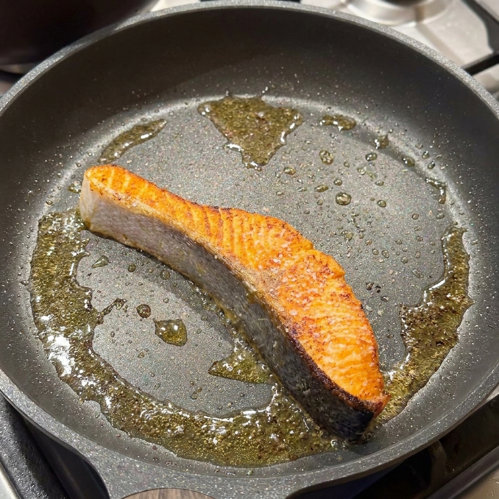 A seared salmon fillet in a frying pan being seasoned with salt and black pepper.