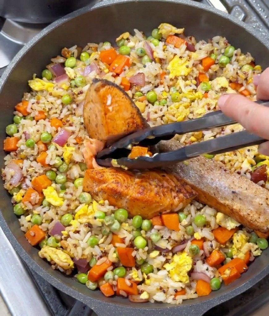 Black kitchen tongs holding a piece of seared salmon over a pan of fried rice and vegetables.