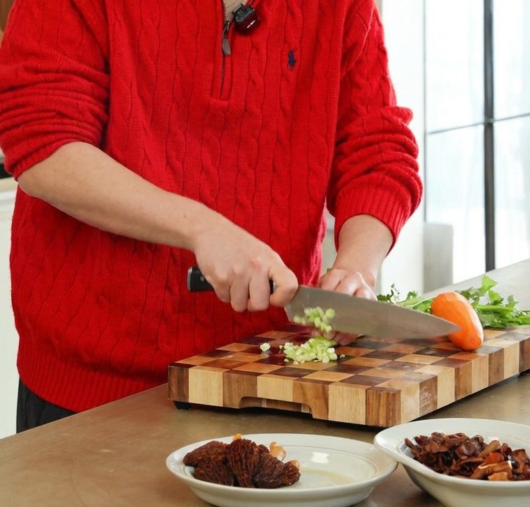 A person using a professional knife to finely dice green celery and orange carrots on a wooden cutting board.