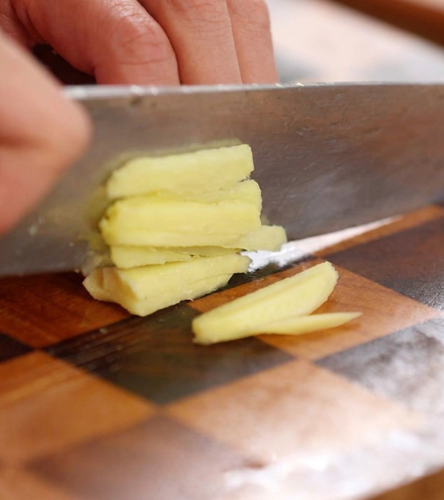 Close-up of a knife finely mincing a slice of fresh ginger into tiny pieces on a wooden board.