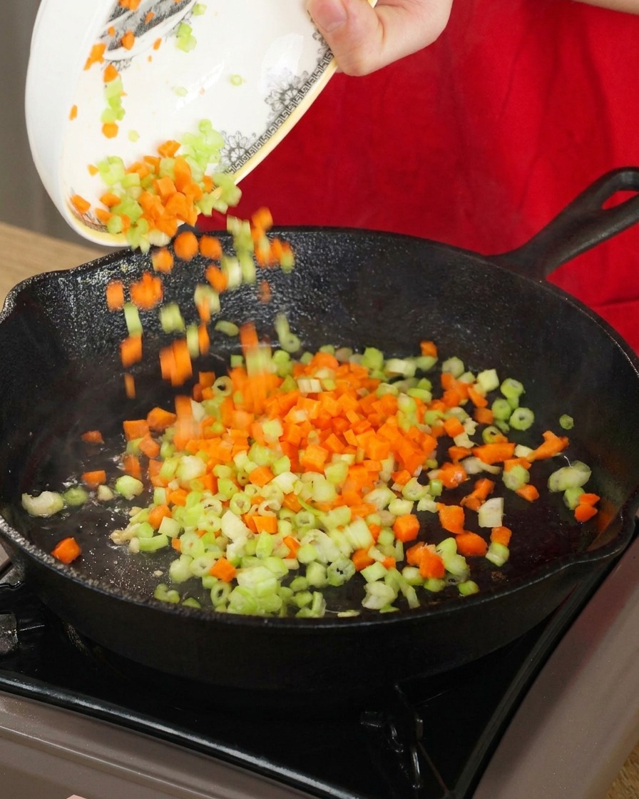 Diced orange carrots and green celery being poured from a decorated white bowl into a hot black cast-iron skillet for stir-frying.