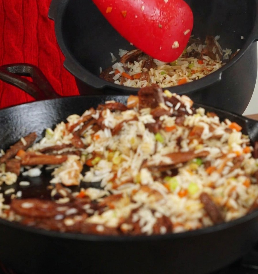 Using a red spatula to move stir-fried rice and mushrooms from a pan into a black rice cooker inner pot.