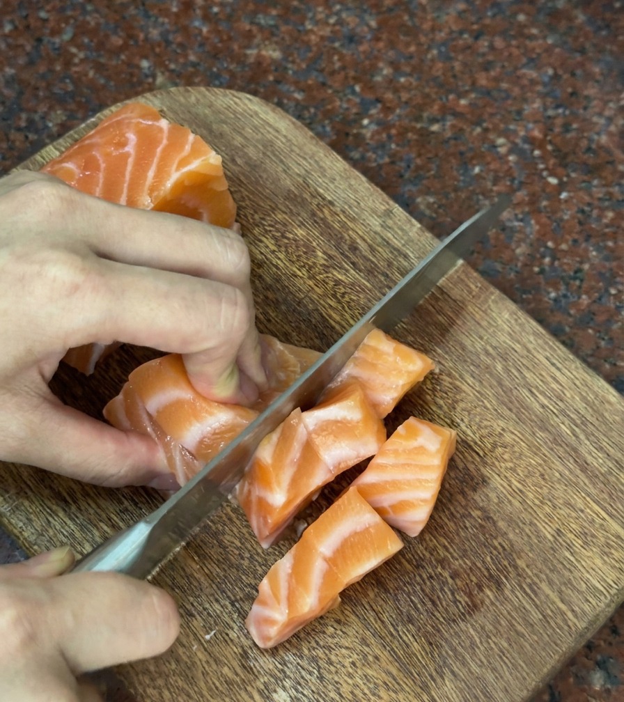 Hands carefully slicing a fresh piece of orange salmon into thick, square cubes on a wooden board.