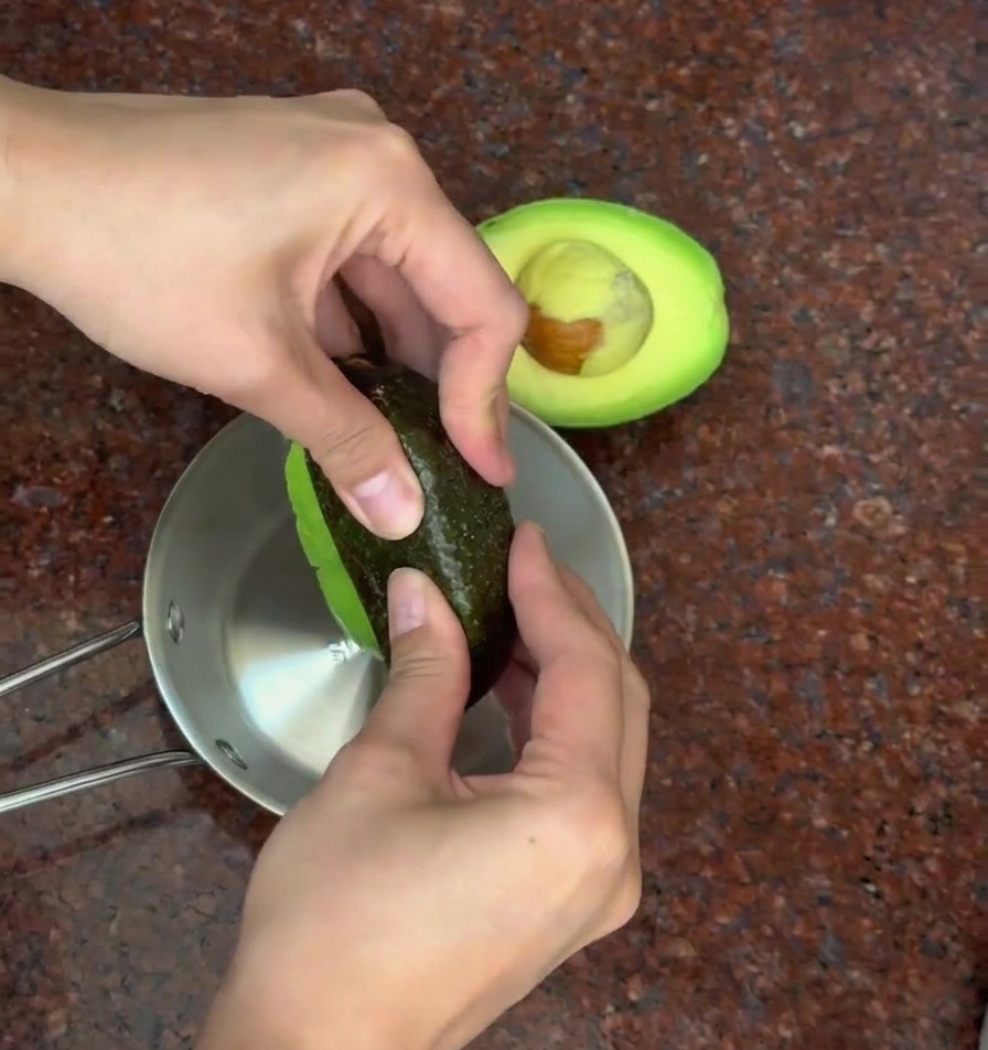 A person using their hands to peel the dark skin away from the green flesh of a halved avocado over a metal bowl.
