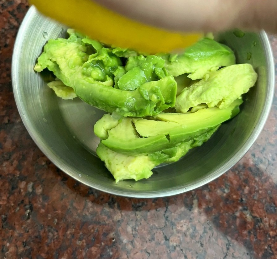 A hand squeezing a yellow lemon over chunks of avocado in a stainless steel bowl.