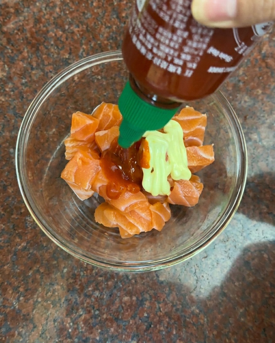 Adding a drizzle of creamy mayonnaise and bright red chili sauce to a bowl of raw salmon cubes.