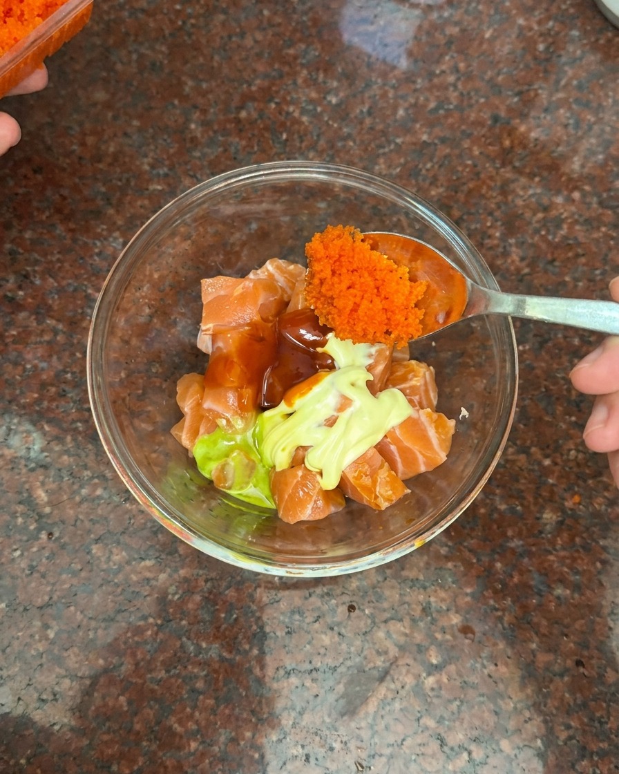 A spoon adding a large portion of tiny orange fish roe to a bowl containing salmon, mayonnaise, and chili sauce.