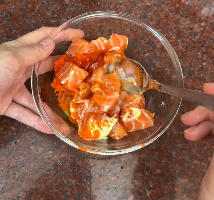 A glass bowl containing fresh salmon cubes being mixed with orange fish roe and a creamy orange sauce using a metal spoon.