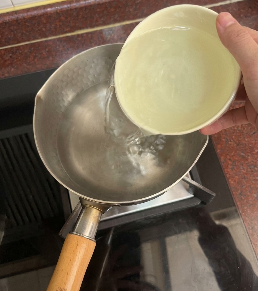 A hand pouring clear water from a light-colored ceramic bowl into a metal pot on a stovetop.