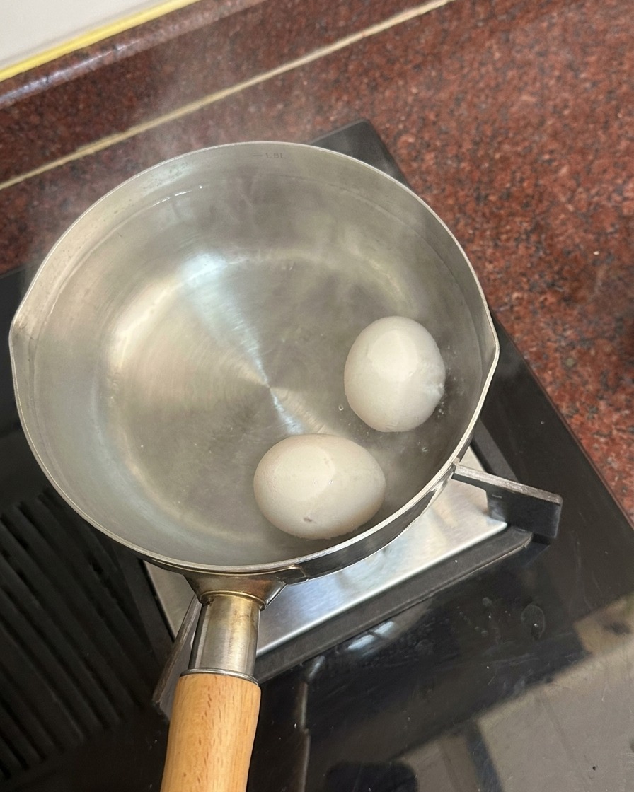 Two white eggs resting at the bottom of a pot filled with hot water, with steam rising from the surface.