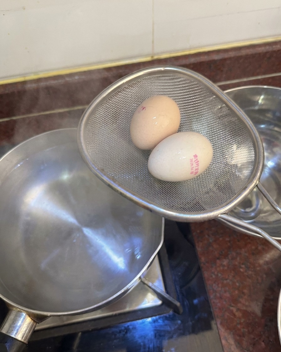 A metal strainer holding two boiled eggs over a pot of water, ready to be placed into a nearby bowl of ice water.