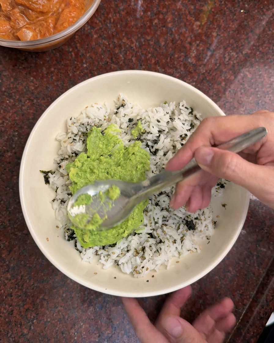 A spoon spreading bright green mashed avocado over a bed of white rice and dark green seaweed flakes in a white bowl.