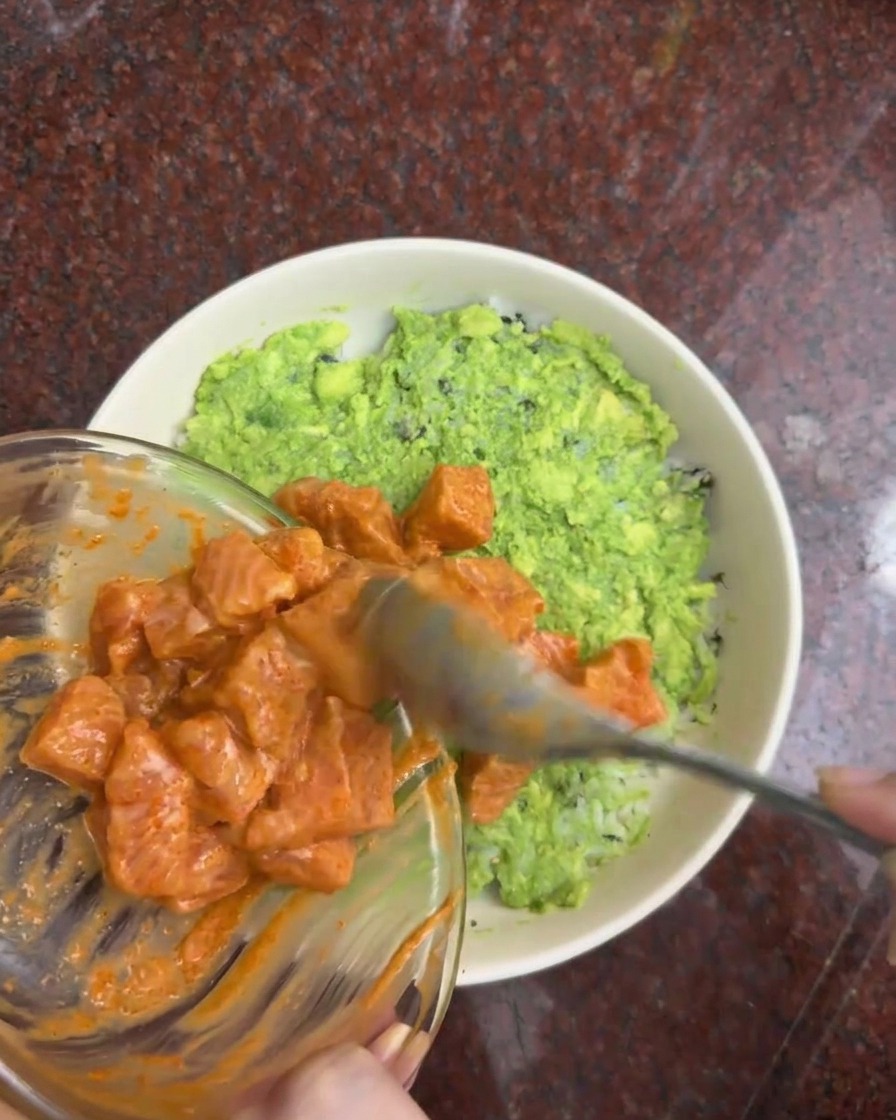 Creamy marinated salmon cubes being transferred from a glass bowl onto a layer of green avocado in a white serving bowl.