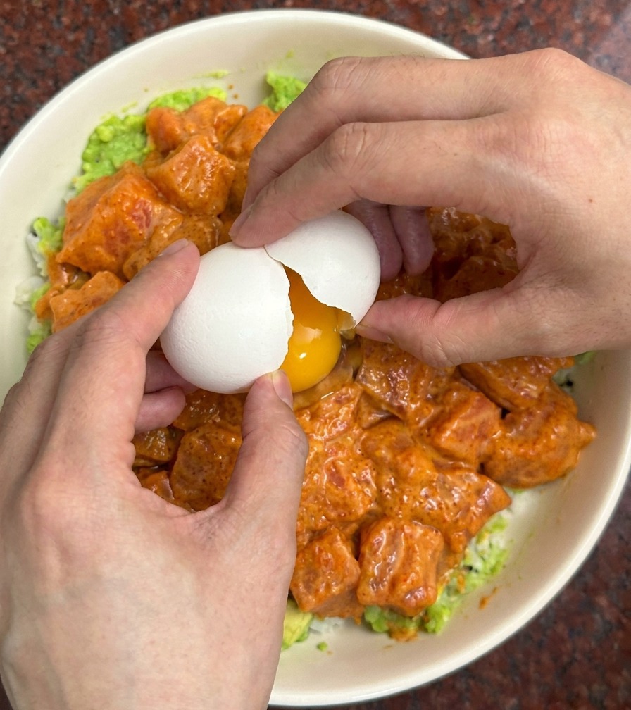 Two hands cracking a white eggshell over a colorful bowl filled with marinated salmon cubes and mashed avocado.