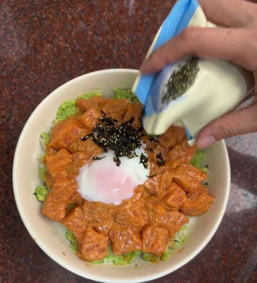 A hand pouring a mixture of black seaweed strips and sesame seeds from a blue and white pouch onto the salmon bowl.
