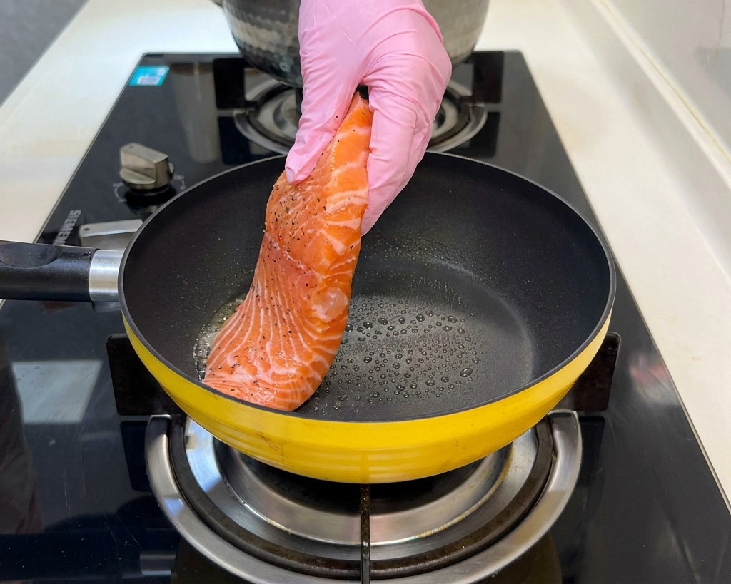 A seasoned raw salmon fillet being placed into a heated yellow frying pan by a hand wearing a pink glove.