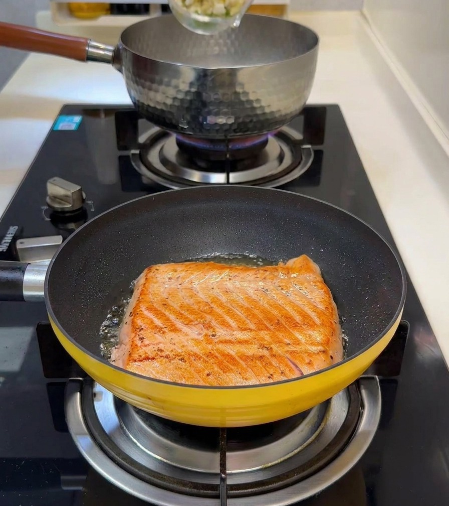 Diced bamboo shoots and green peas being poured into a small pot of boiling water while a salmon fillet sears in a pan in the foreground.