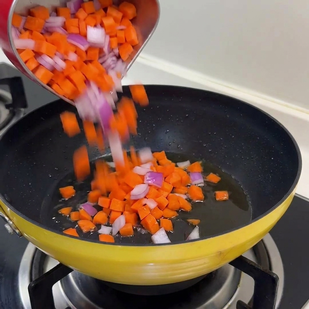 A mixture of diced orange carrots and purple onions being poured into a yellow frying pan with shimmering oil.