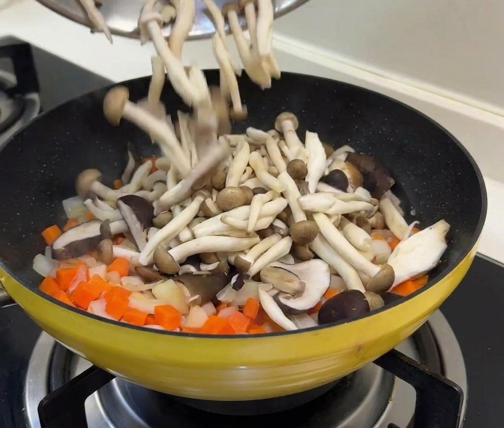 Fresh mushrooms and green peas being poured from a bowl into a pan containing sautéed carrots and onions.