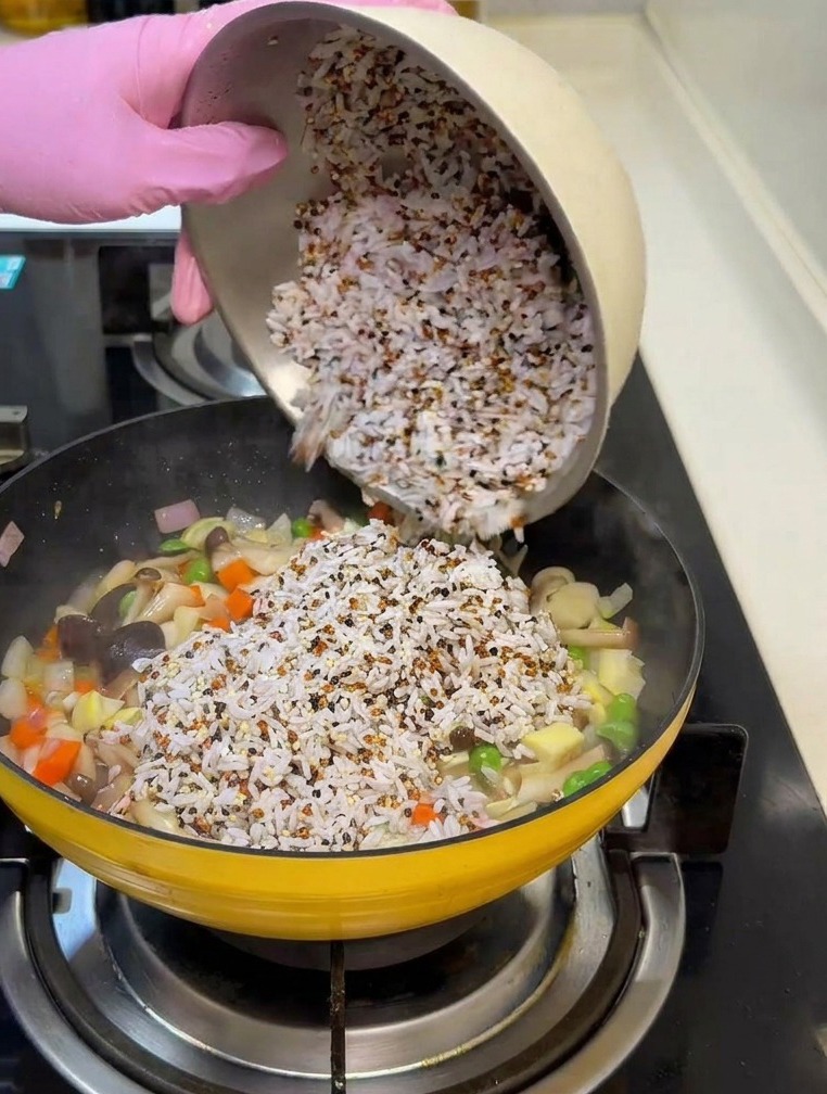 A hand in a pink glove pouring a bowl of mixed white rice and quinoa into a yellow pan containing stir-fried carrots, onions, and mushrooms.