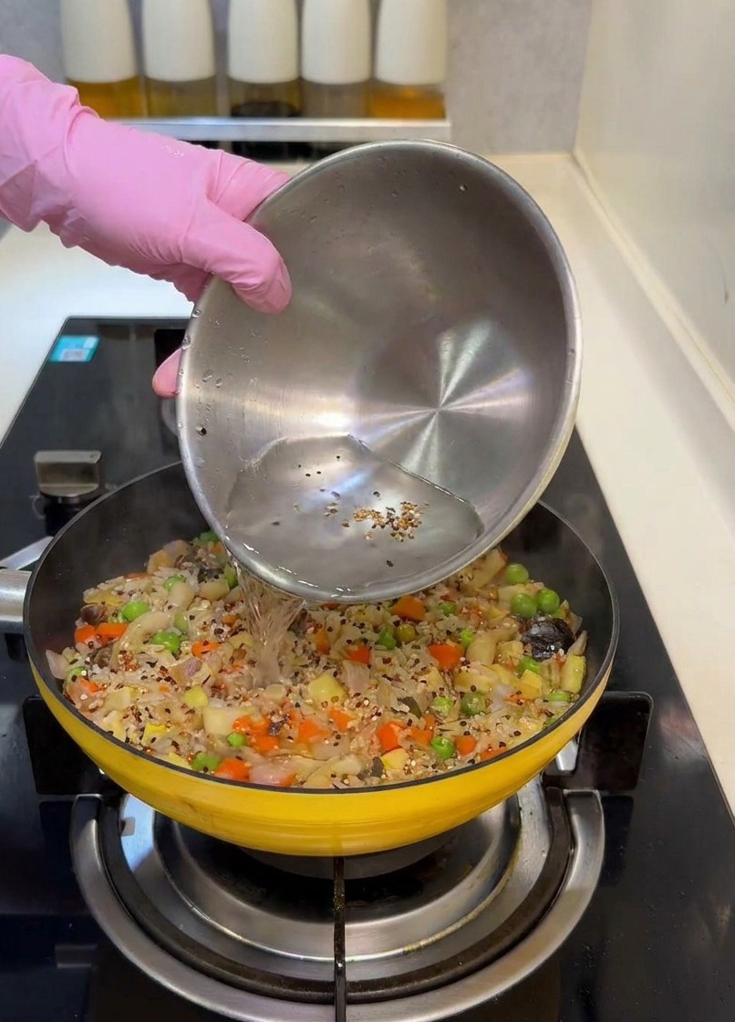 Water being poured from a stainless steel bowl into a yellow pan filled with a colorful rice and vegetable mixture.