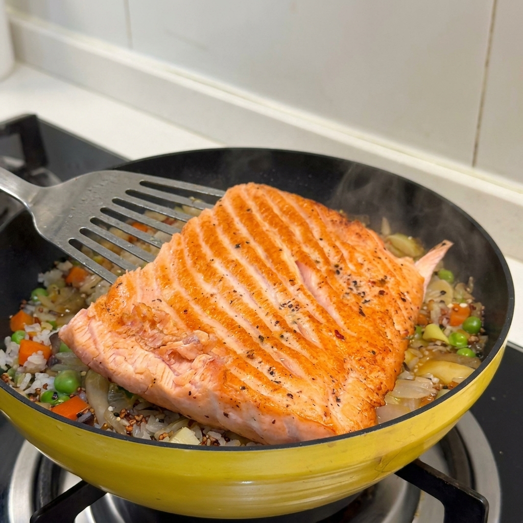 A large, golden-brown seared salmon fillet being lowered onto a bed of seasoned rice and mixed vegetables in a yellow pan.