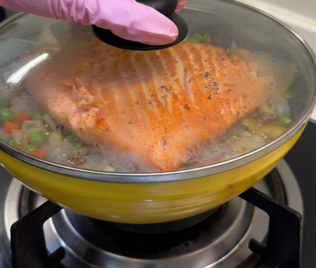 A yellow pan containing salmon and vegetable rice being covered with a clear glass lid to trap steam.