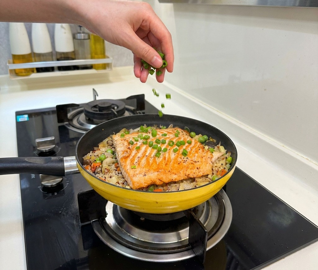 A hand sprinkling bright green chopped onions over a finished salmon and vegetable braised rice dish.