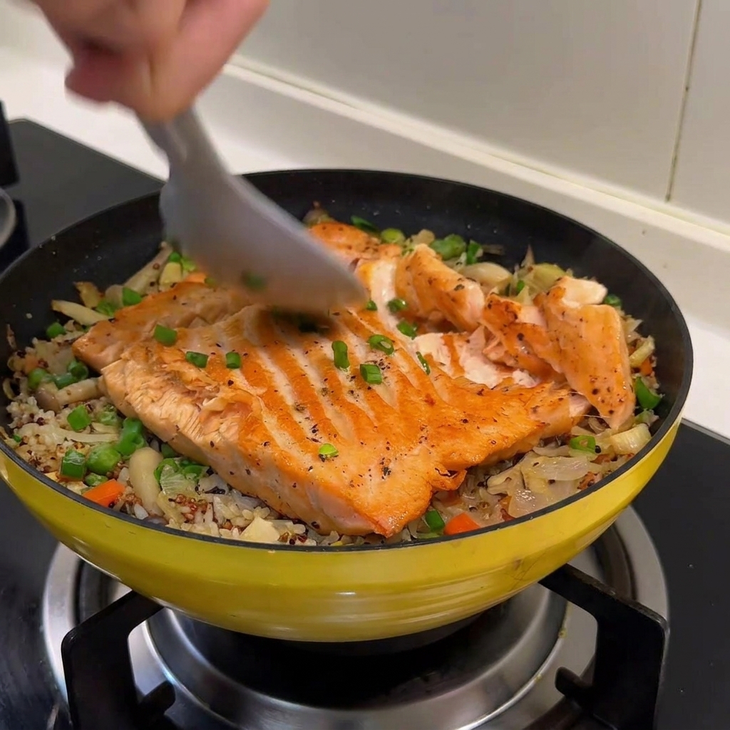 A metal spoon breaking a cooked salmon fillet into flakes on top of a bed of braised rice and spring vegetables.