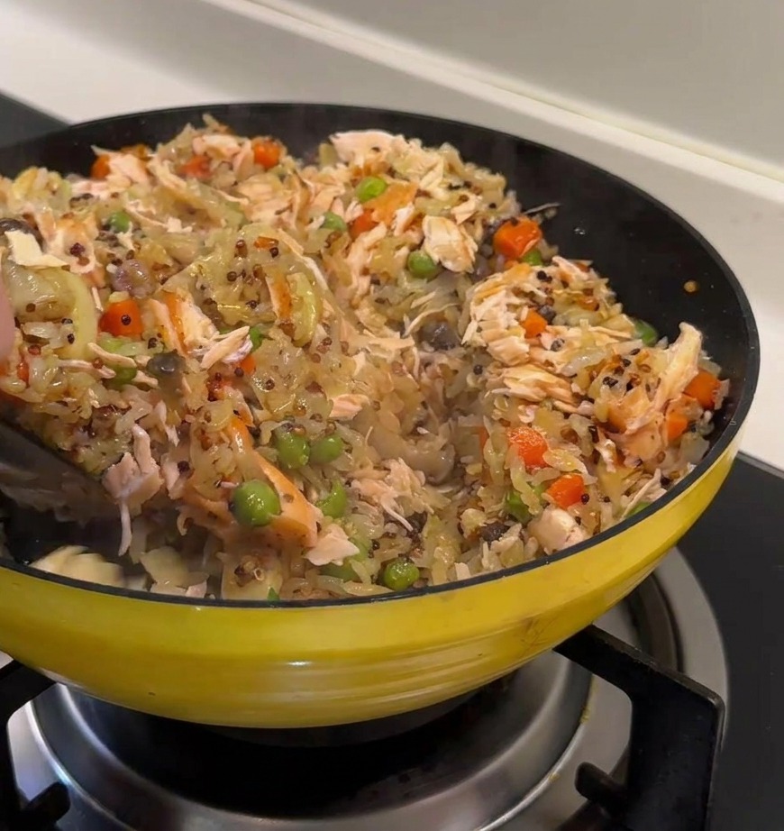 A hand using a black spatula to fold large flakes of cooked salmon into a colorful pan of quinoa rice, peas, and diced carrots.