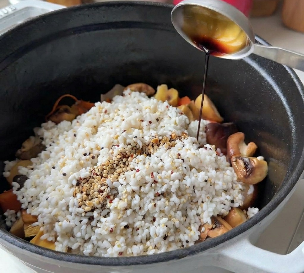 A metal spoon pouring dark soy sauce over a mound of white rice and quinoa mixed with sliced mushrooms and vegetables in a black pot.