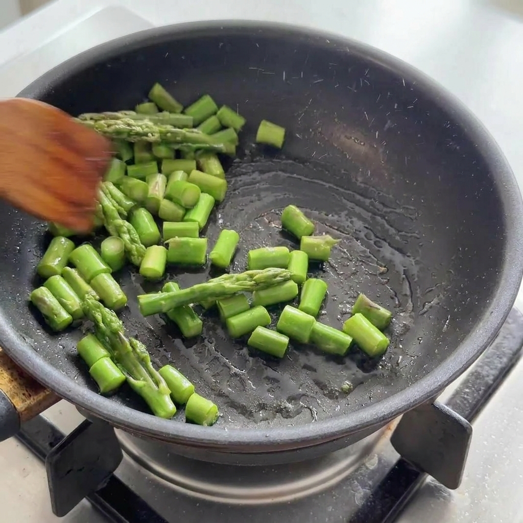 Green asparagus sections being tossed in a black frying pan with a wooden spatula.