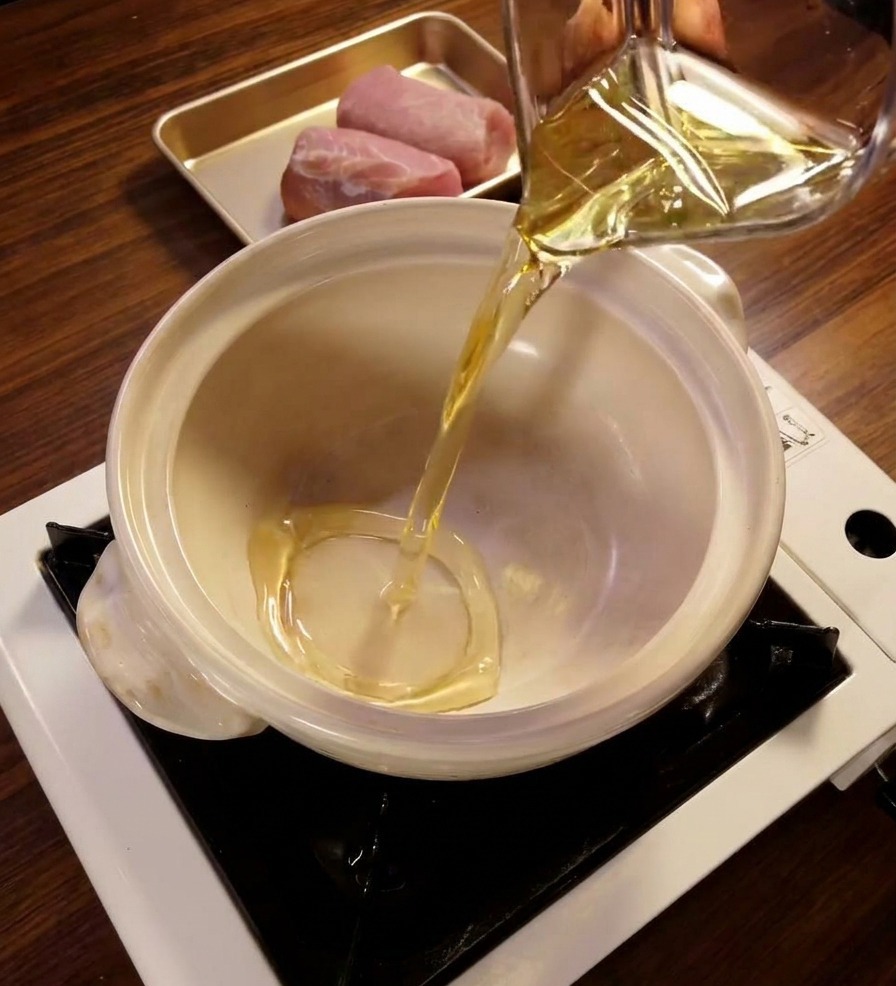 Clear mirin liquid being poured from a glass measuring cup into a clean white ceramic pot on a portable stove