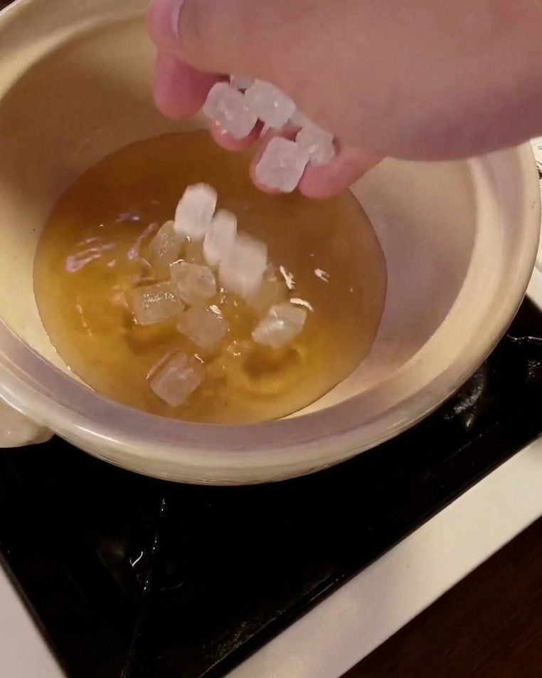 A hand dropping translucent rock sugar crystals into a ceramic pot containing mirin liquid.