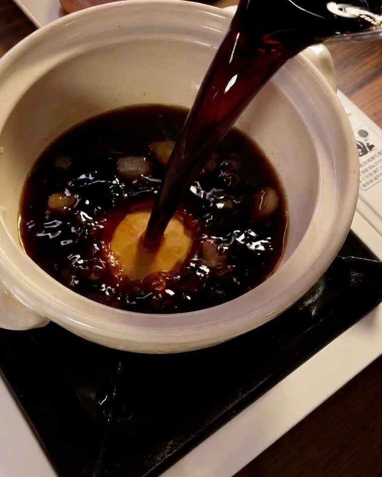 Dark soy sauce being poured into a ceramic pot containing mirin and rock sugar crystals.